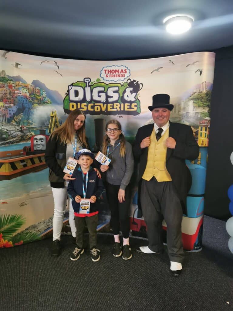 Three children and one adult man dressed as the Fat Controller pose in front of a Thomas & Friends: Digs & Discoveries backdrop. One child holds a Thomas toy, and all three children have lanyards. The background features colourful scenery and Thomas the Tank Engine.