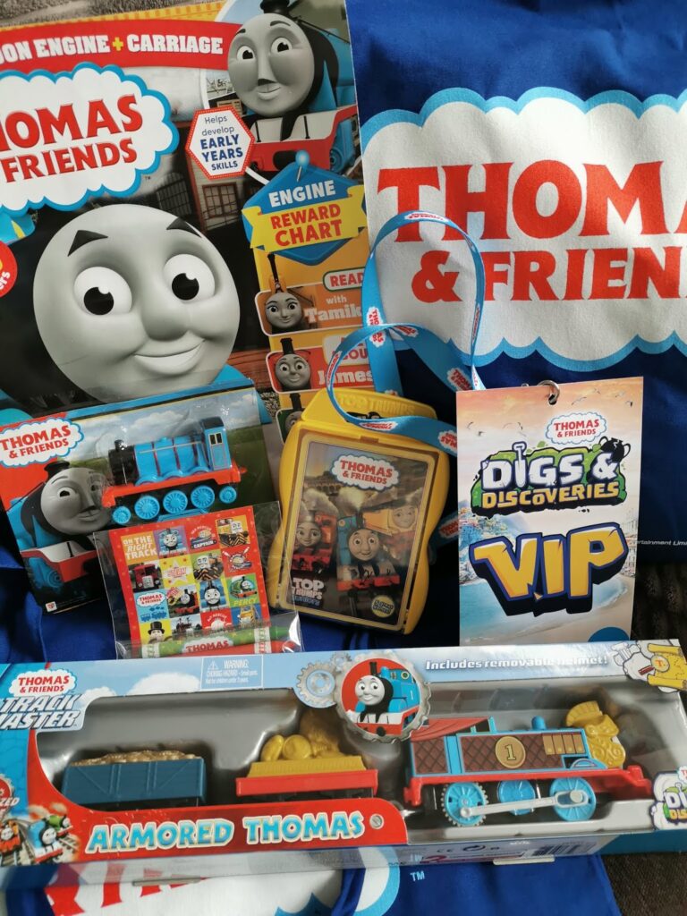 A collection of Thomas & Friends themed items, including a magazine, sticker sheets, a Thomas toy train set, a blue bag, a yellow lunchbox, a blue lanyard, a VIP badge, and a boxed “Armoured Thomas” train toy. Bright, colourful designs throughout.