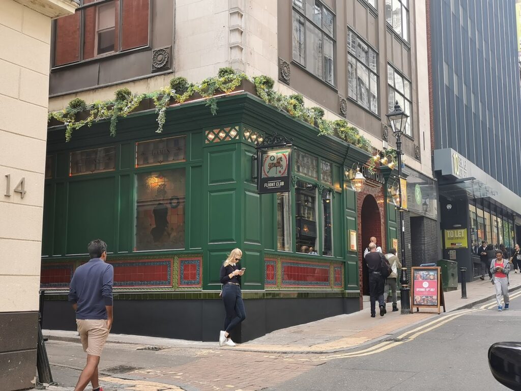 A green pub with red tile details and hanging plants is on a street corner. People walk by, including a man in a blue jacket and a woman on her mobile. A black sign reads "The Punch Bowl." Tall buildings line the narrow, slightly curved street.