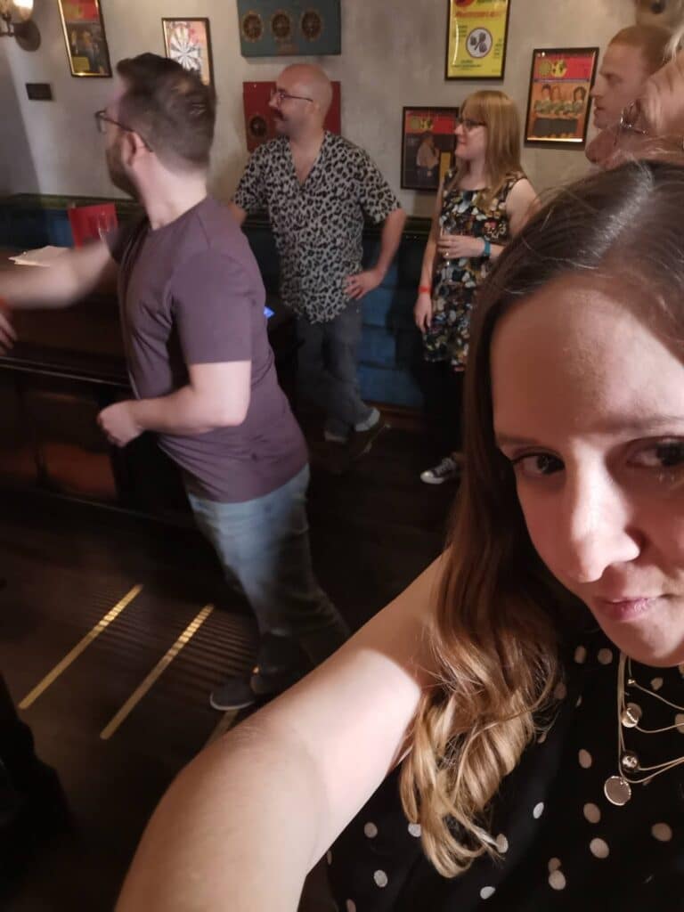 A woman in a black polka dot top takes a selfie in a bar with four people in the background. One man is throwing a dart, whilst others watch. The room has framed pictures, dartboards, and dim lighting, creating a lively, casual atmosphere.