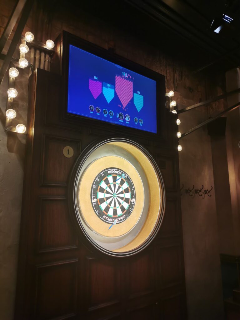 A dartboard is mounted on a wooden wall with a large circular frame and lit by surrounding bulbs. Above the dartboard, a digital screen displays a game interface with scores and coloured sections. The setting appears to be inside a bar or games room.