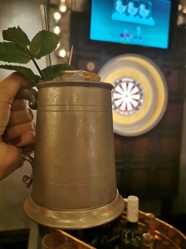 A hand holds up a metallic tankard garnished with mint leaves in front of a dartboard. In the background, a digital screen displays a darts game, and bottles sit on a table below the dartboard. The scene appears to be in a bar or pub setting.