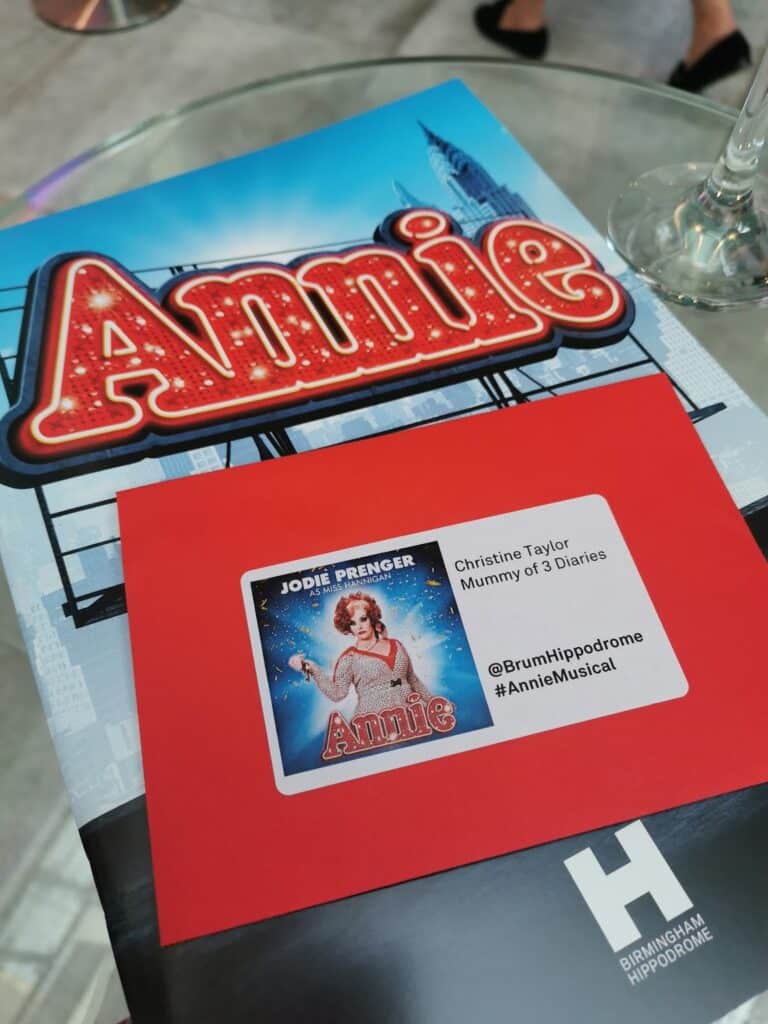 A red envelope labelled with “Jodie Prenger Annie” and event details rests on a glossy Annie musical programme. The programme features the illuminated Annie logo. The envelope partially covers the programme, and both items are on a round glass table.