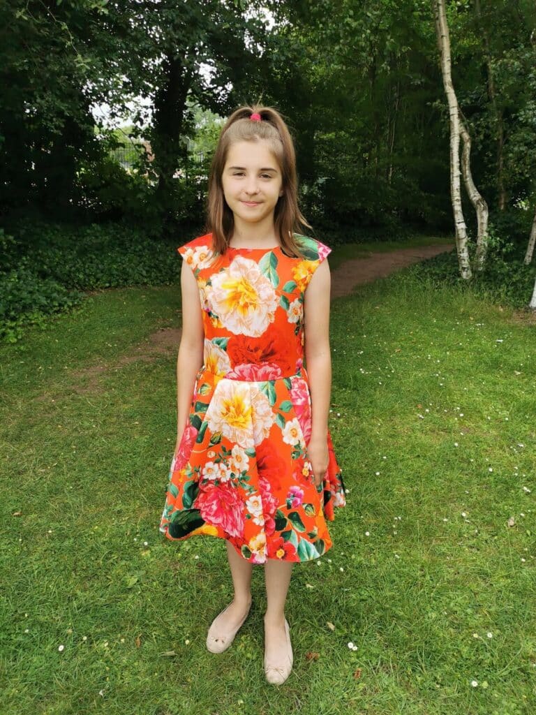 A young girl with light brown hair in a half-up style stands on grass in a park, wearing a bright orange dress with large, colourful floral patterns. She is smiling softly and has beige shoes. Lush green trees and bushes are in the background.