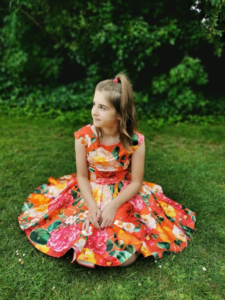 A young girl with light brown hair in a ponytail sits cross-legged on green grass, wearing a vibrant, floral-patterned dress in shades of orange, pink, yellow, and green. She looks to her left, with lush green bushes in the background.