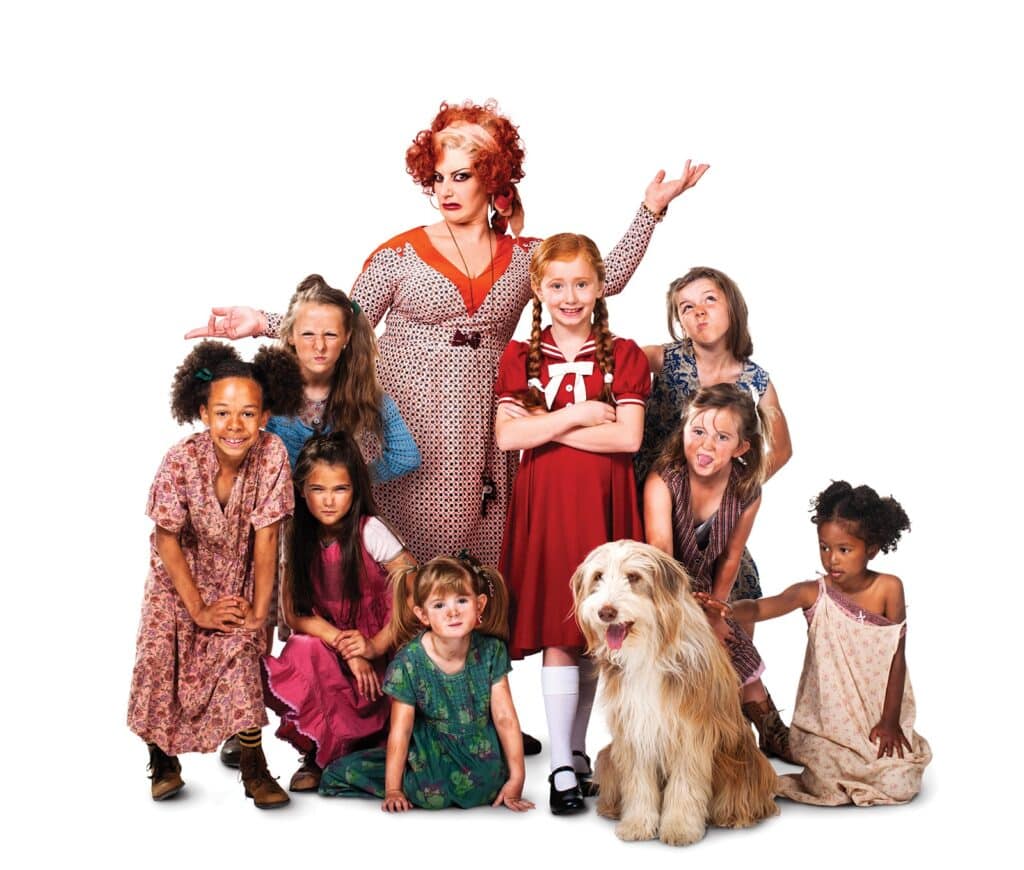 A group of young girls in worn dresses pose with a woman in a red and white dress, who stands with a stern expression. A cheerful girl in a red dress and white socks stands in front of her. A fluffy dog sits at the group's feet. The background is plain white.
