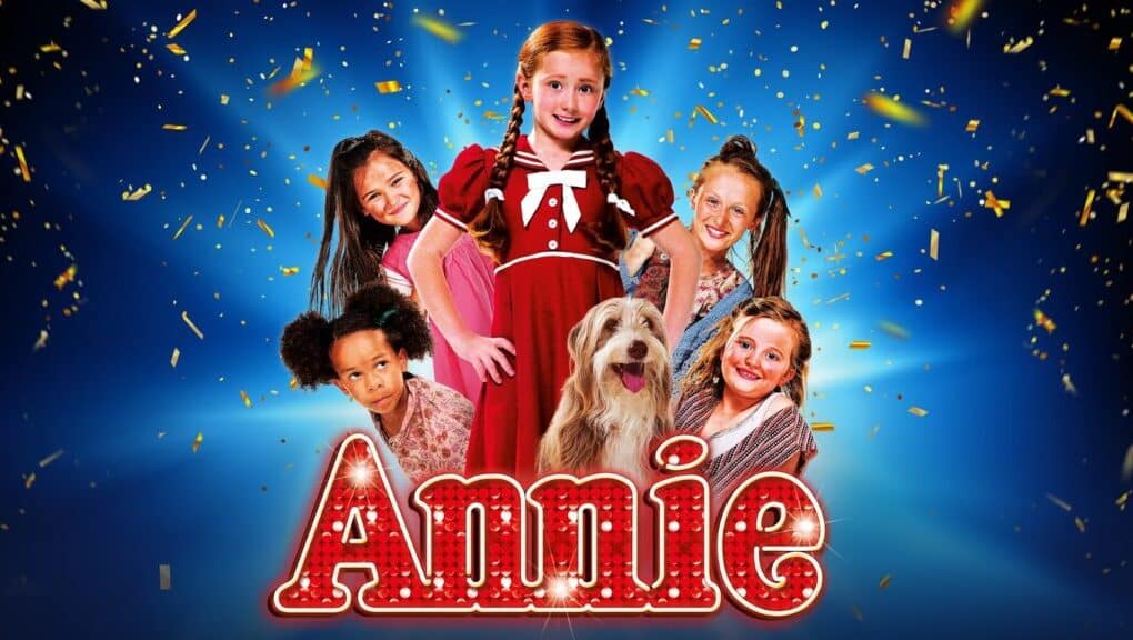 A group of smiling children in colourful clothes pose with a shaggy dog in front of a blue background with gold confetti. The girl in the centre wears a red dress and pigtails. Large, lit-up letters spell "Annie" at the bottom.