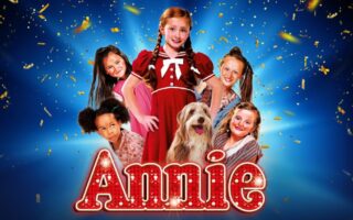 A group of smiling children in colourful clothes pose with a shaggy dog in front of a blue background with gold confetti. The girl in the centre wears a red dress and pigtails. Large, lit-up letters spell "Annie" at the bottom.