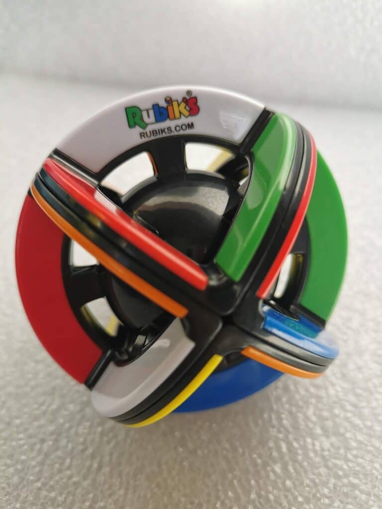 A spherical Rubik’s puzzle with intersecting coloured bands—red, green, blue, white, yellow, and orange—on a glossy black base. The Rubik’s logo and website appear at the top. The puzzle is photographed against a textured, light grey background.