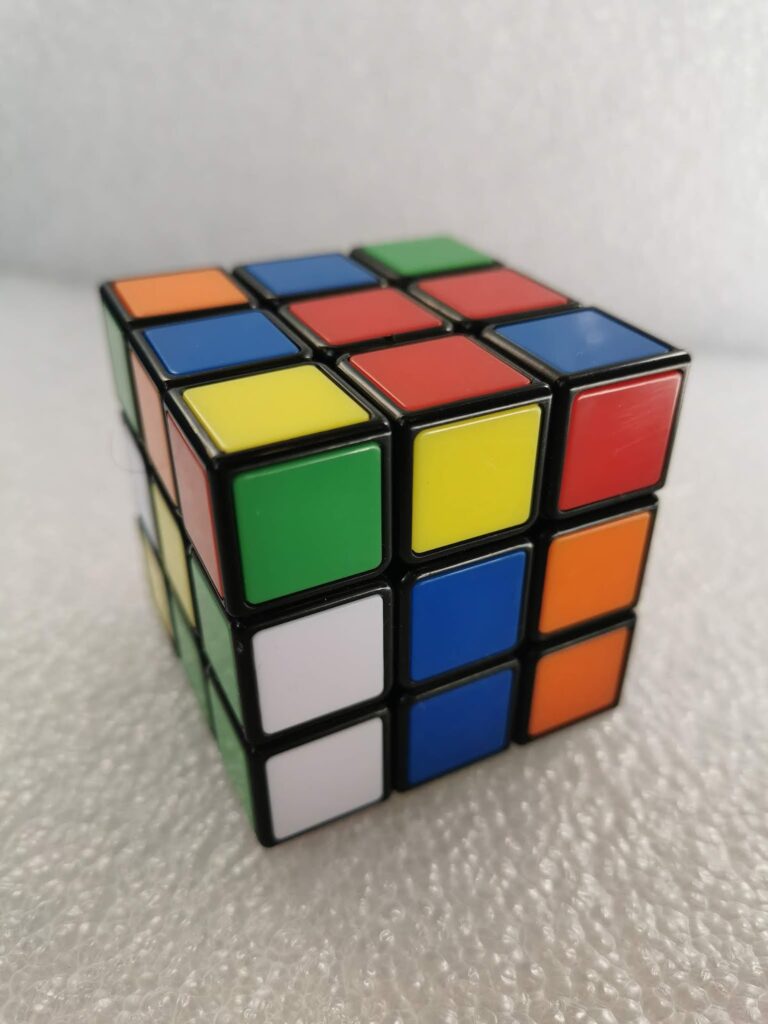 A partially solved Rubik’s Cube sits on a textured white surface. Its visible sides show mixed coloured squares—blue, red, yellow, green, orange, and white—none of which are fully solved. The black frame separates each coloured tile.