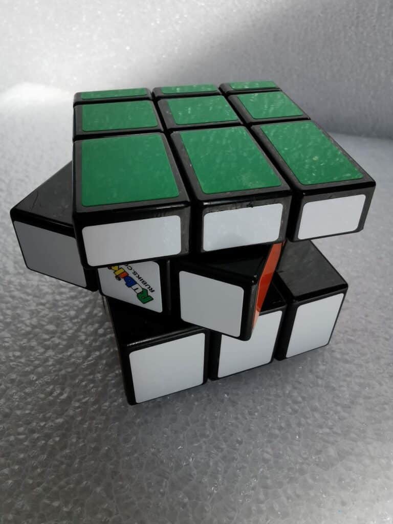 A partially twisted Rubik’s Cube rests on a textured, bubble wrap surface. The visible faces show green and white stickers, with one orange sticker partially visible. The cube is misaligned, creating an irregular, staggered appearance.