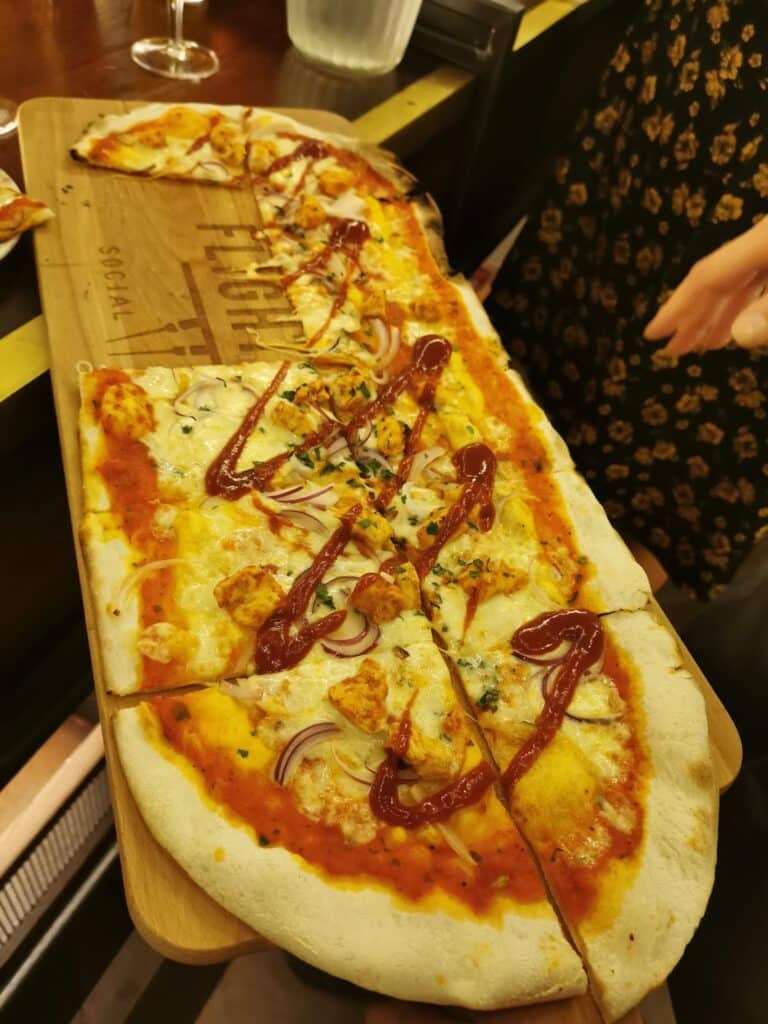 A rectangular flatbread pizza is served on a wooden board. It is topped with melted cheese, thin red onion slices, herbs, and drizzled with a sauce similar to tomato ketchup. A hand reaches for a slice. Part of a floral dress is visible. Glasses and a jug are in the background.