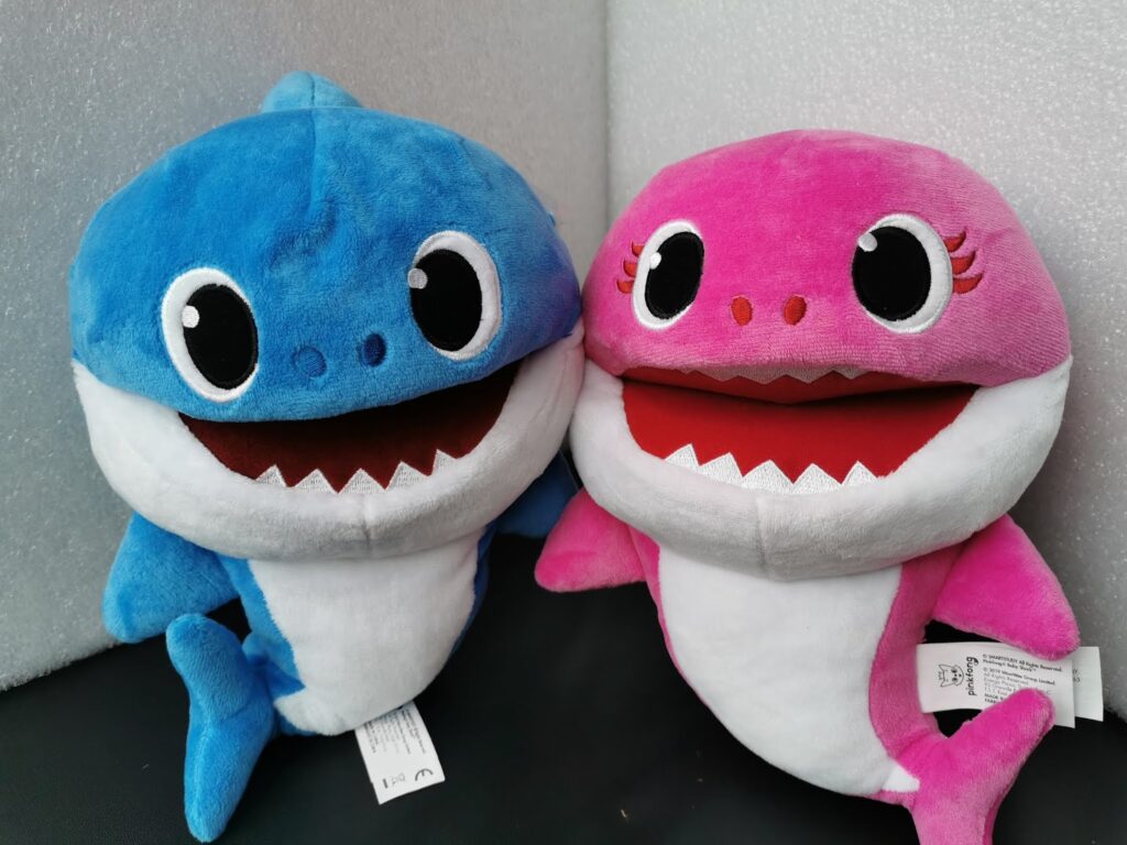 Two plush toy sharks sit side by side—the left one is blue with large black and white eyes, and the right one is pink with matching eyes, eyelashes, and rosy cheeks. Both have wide, open mouths showing white felt teeth and white bellies.