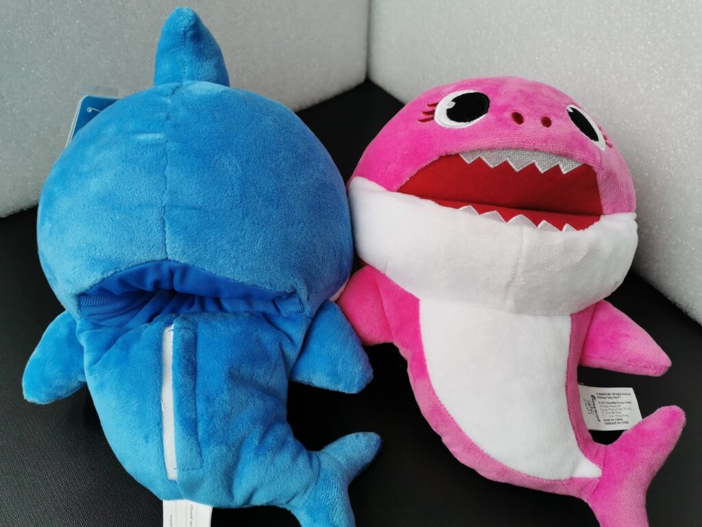 Two plush shark toys sit on a black surface. The left one is blue with a zip on its back, facing away. The right one is pink with big eyes, an open mouth showing white teeth, and a white belly, facing forward. Both have tags attached.