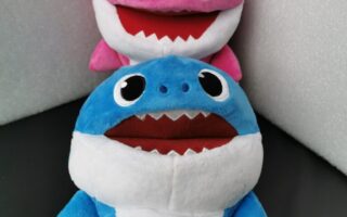 Two plush toy sharks are shown. The front toy is blue with a white belly and wide open mouth, while the one behind is pink with similar features. Both have big, round eyes and are positioned against a black and white background. A tag is attached to the pink shark.