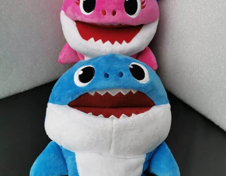 Two plush toy sharks are shown. The front toy is blue with a white belly and wide open mouth, while the one behind is pink with similar features. Both have big, round eyes and are positioned against a black and white background. A tag is attached to the pink shark.