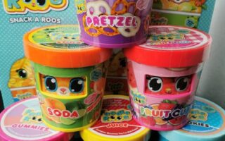 Six colourful Foodie Roos toy containers are stacked, each with cute animal faces and snack themes: Pretzel, Soda, Fruit Cup, Gummies, Cookies, and Juice. A Foodie Roos Snack A Roos box with a cartoon tiger design is in the background.