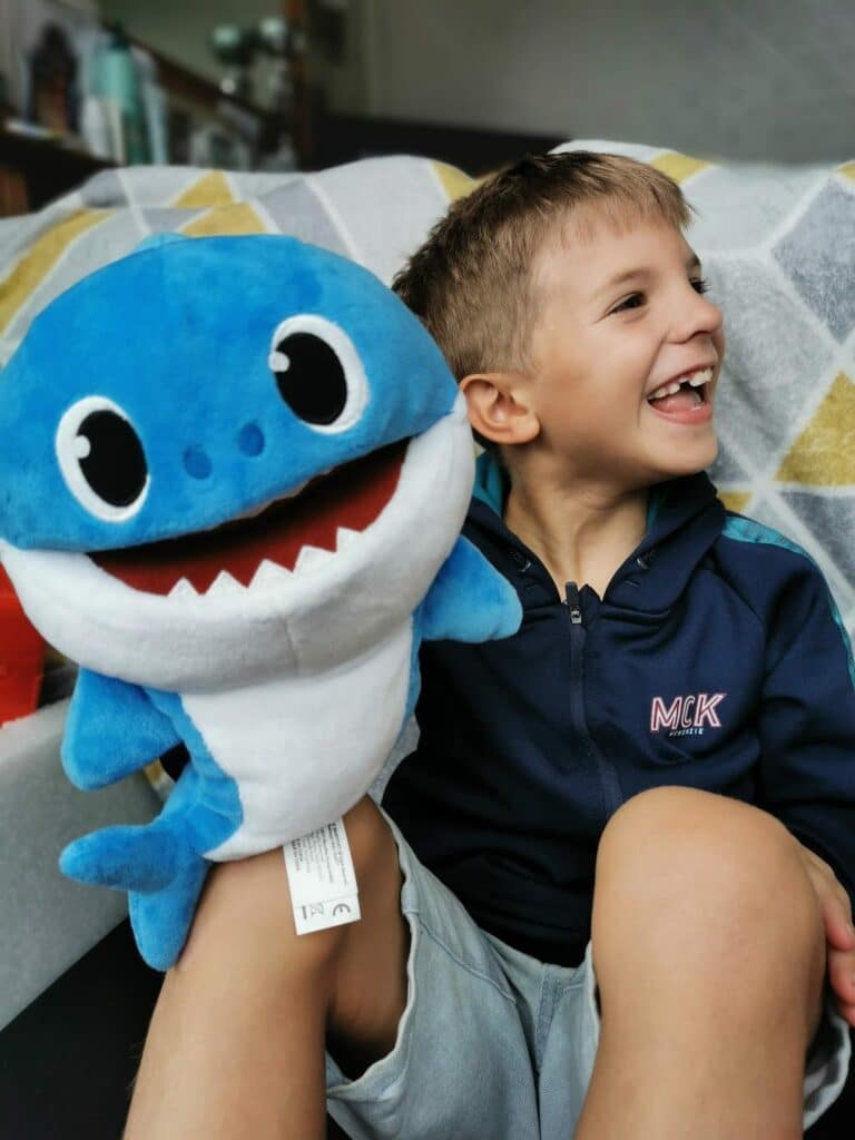 A young boy with short blond hair, wearing a navy jacket and grey shorts, sits on a sofa smiling widely. He holds a large, blue cuddly toy shark with big eyes and an open mouth. The background shows a patterned blanket draped over the sofa.