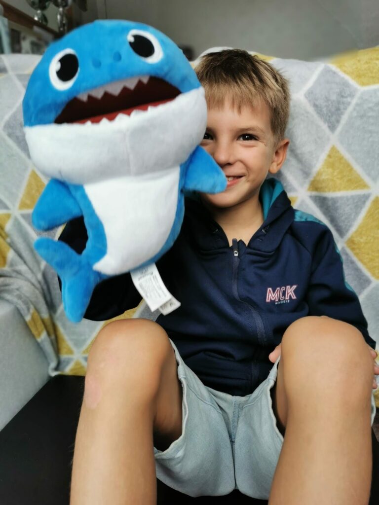 A young boy with short blond hair sits on a sofa, smiling and holding up a blue and white plush shark toy. He is wearing a navy hoodie and light grey shorts. The background features a grey and yellow geometric patterned blanket.