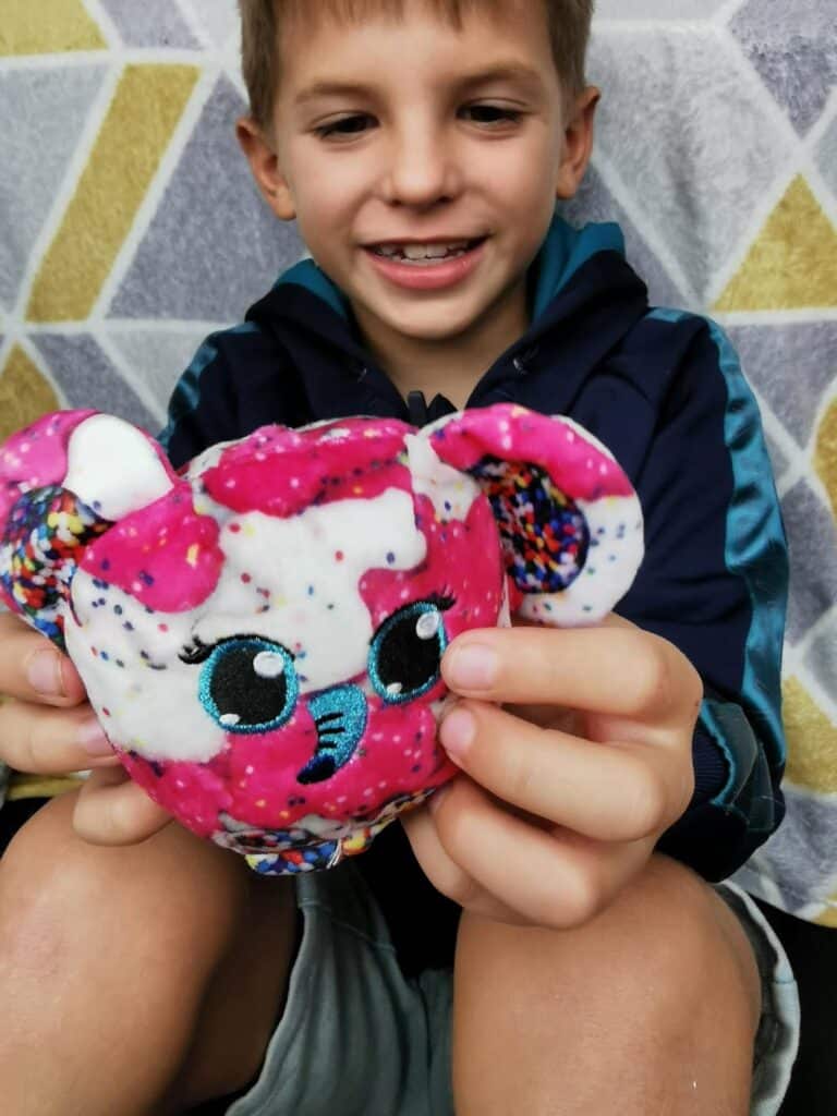 A smiling young boy in a navy jacket holds a pink soft toy with big embroidered eyes and sprinkle-like patterns on its ears and head. The background shows a geometric-patterned blanket in white, yellow, and grey tones.
