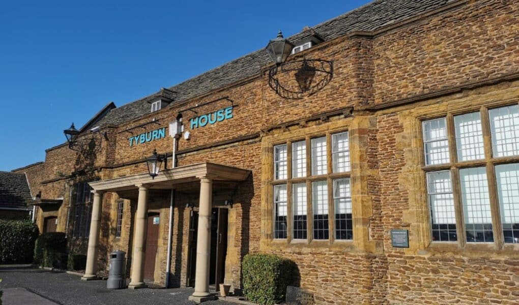 A brown stone building with large grid-paned windows and a portico entrance. The sign above the entrance reads “TYBURN HOUSE” in blue letters. There are bushes, two black lamps, and a clear blue sky overhead.