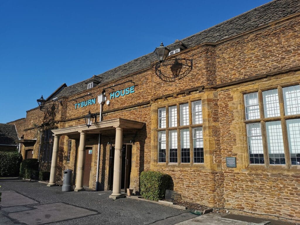 A brown stone building with large rectangular windows, a sloped tiled roof, and "Tyburn House" written in teal letters above the entrance, which is framed by columns and two lanterns. Shrubs line the front, and the sky is bright blue.