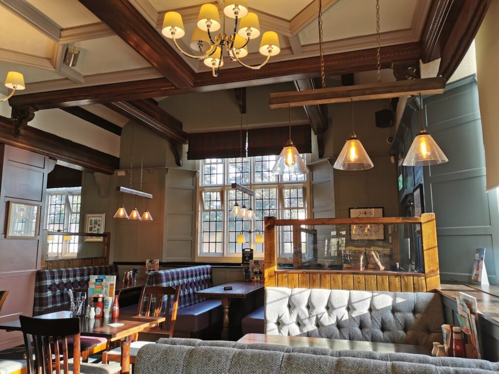 A sunlit pub interior with wooden beams, hanging pendant lights, cushioned booths, and wooden tables. Large windows let in natural light, casting shadows. Menus and condiments sit on tables, and framed art decorates the cream panelled walls.