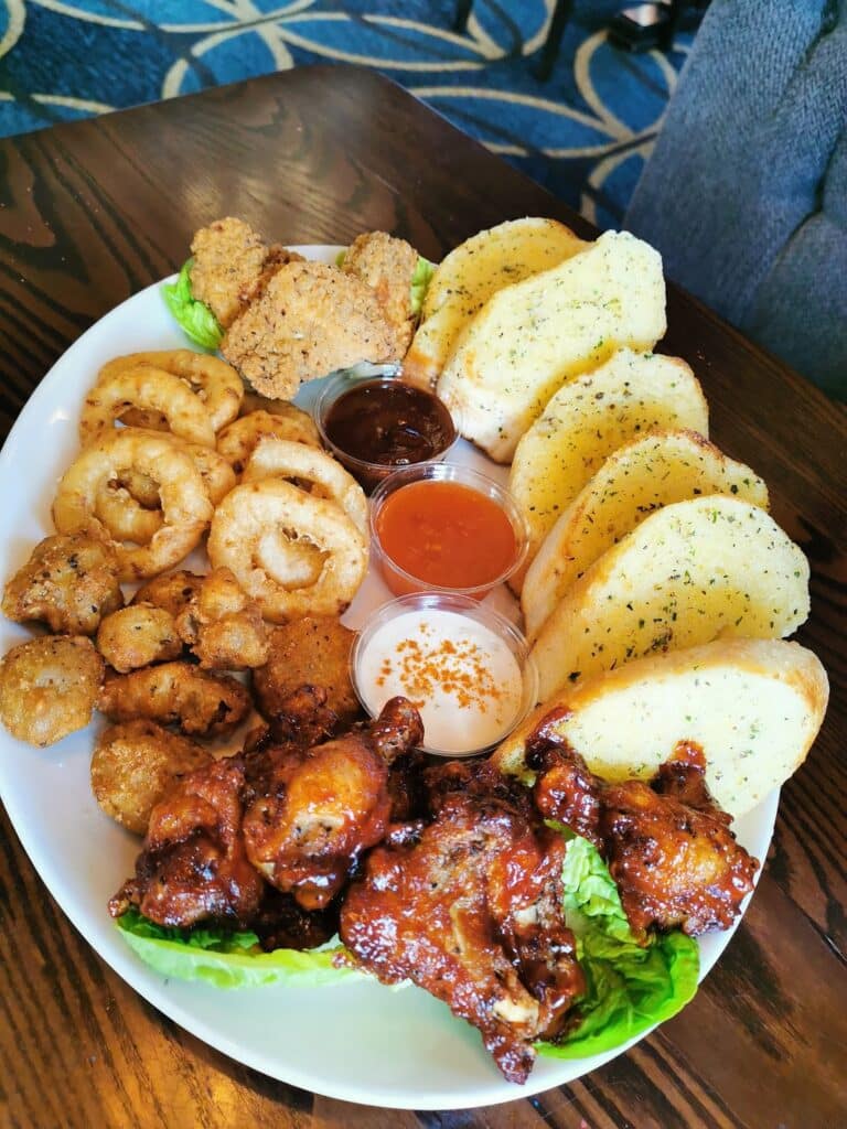A large white plate with garlic bread slices, fried onion rings, breaded mushrooms, two fried chicken goujons, glazed chicken wings, and small pots of barbecue sauce, buffalo sauce, and ranch dressing sits on a wooden table. Blue patterned carpet is visible in the background.