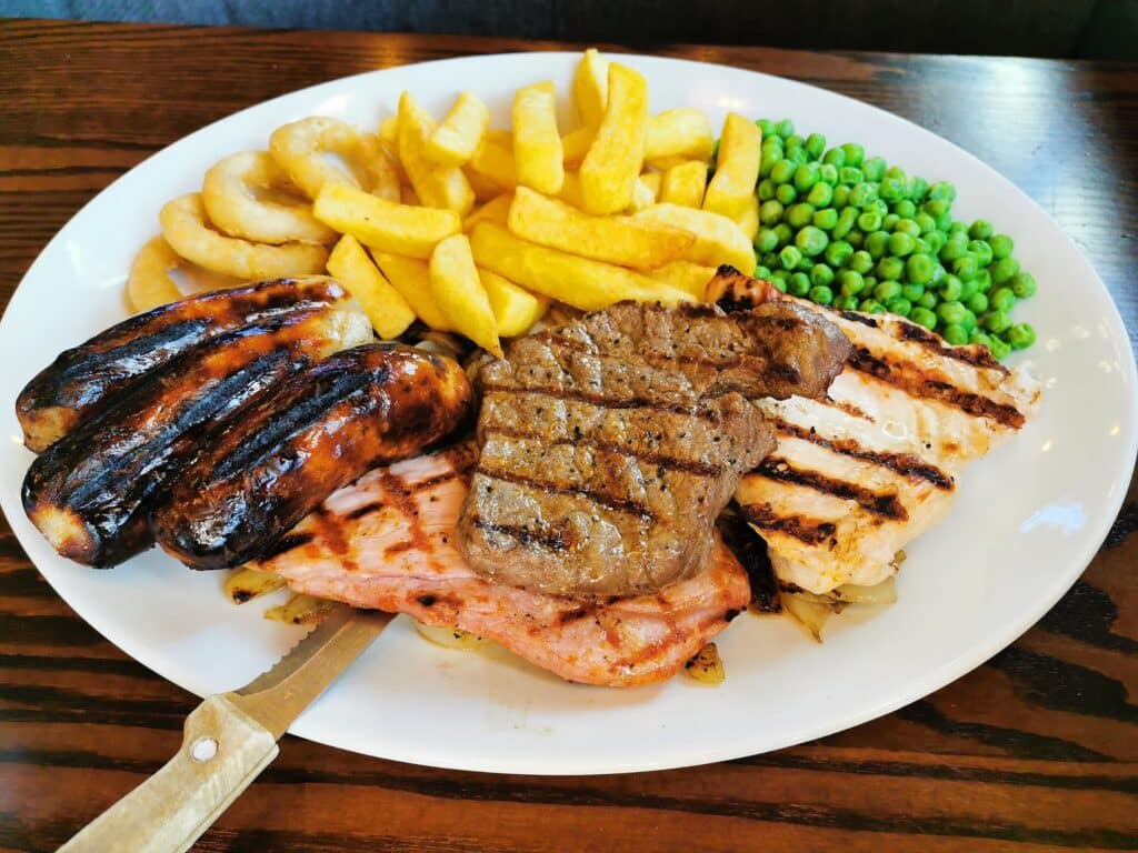 A white plate with grilled sausages, a grilled steak, grilled chicken, thick-cut chips, onion rings, peas, and a slice of gammon on a wooden table. A steak knife rests under the gammon in the bottom left corner of the plate.
