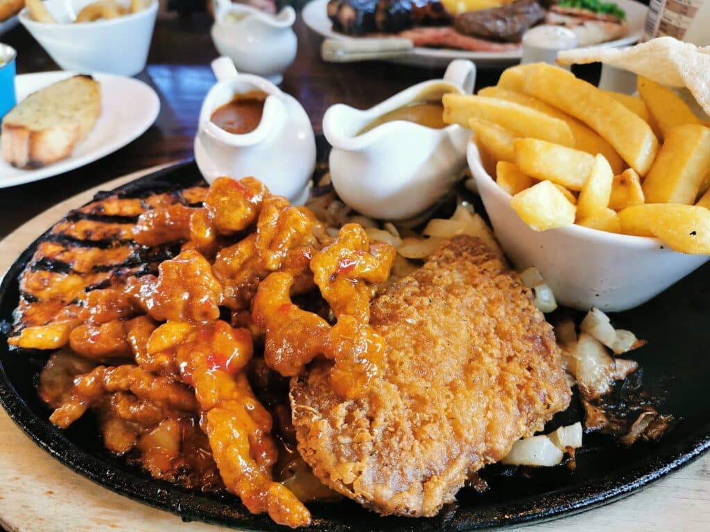 A sizzling platter features grilled chicken strips in a red sauce, a breaded fried escalope, sautéed onions, a bowl of golden chips, and two small white gravy boats, with garlic bread and more food visible in the blurred background.