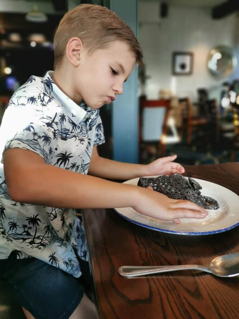 A young boy with short blond hair, wearing a white shirt with a black palm tree pattern and dark shorts, sits at a wooden table. He presses both hands onto a pile of dark, textured food on a white plate, looking focused. A spoon lies nearby.