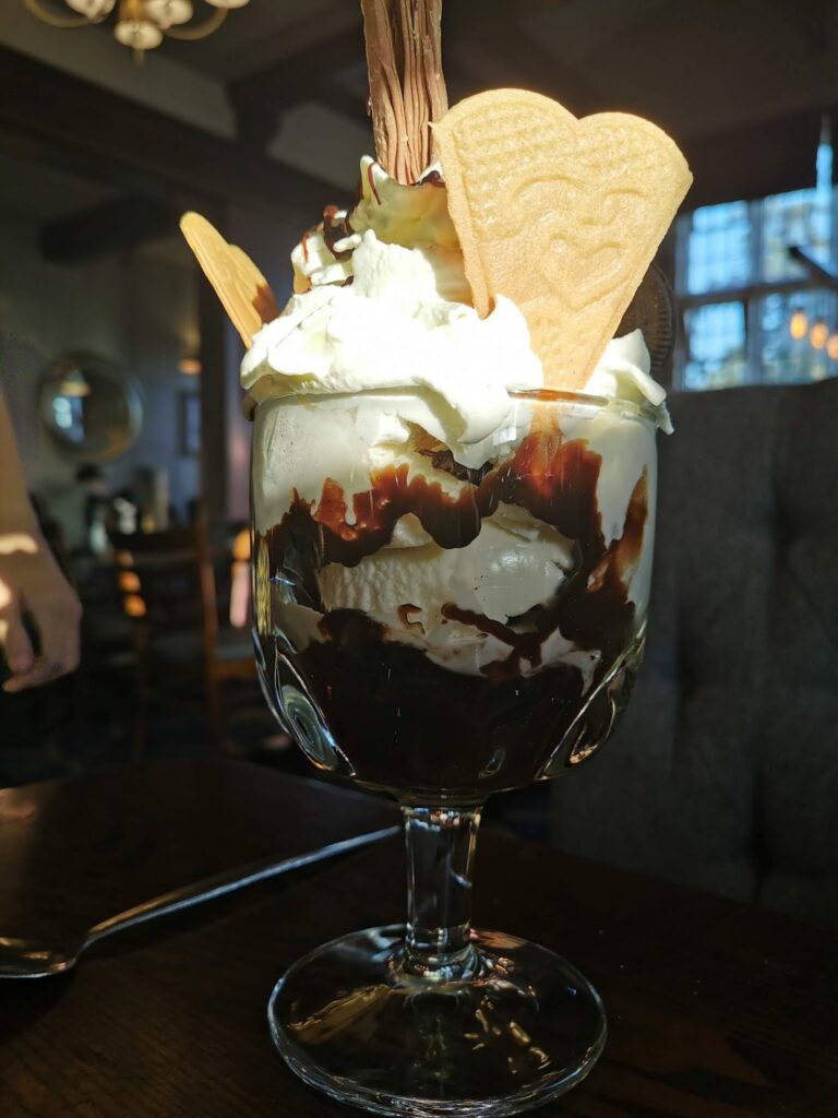 A glass sundae dish filled with vanilla ice cream, whipped cream, chocolate sauce, a chocolate Flake stick, and two heart-shaped biscuits. The dessert is sitting on a dark table in a softly lit restaurant, with blurred chairs and windows in the background.