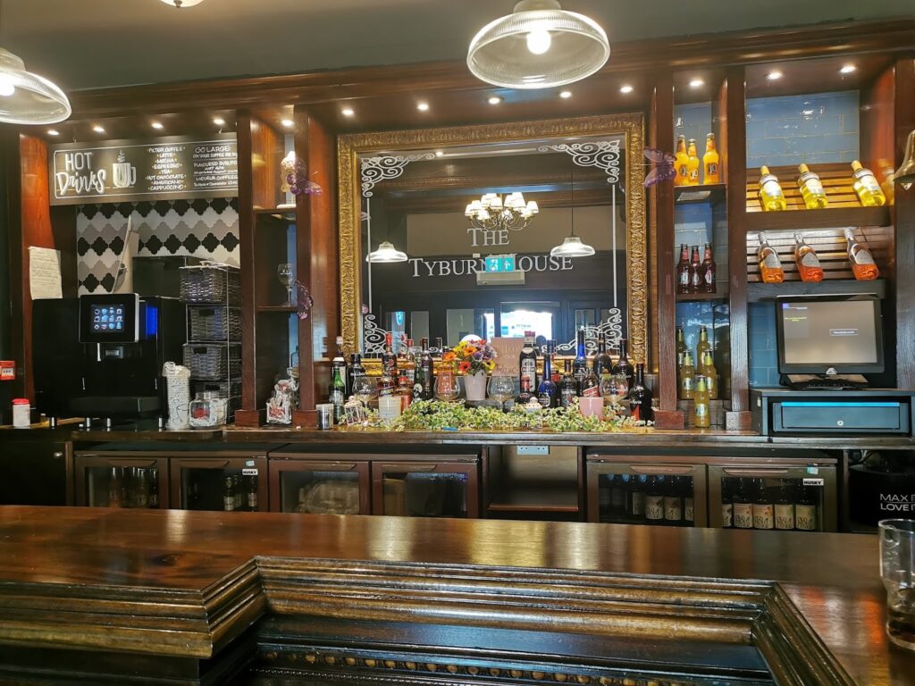A wooden bar counter with various liquor bottles, beer taps, and a bouquet of flowers. Behind the bar, shelves hold more bottles and a coffee machine. A large mirror reads “THE TYBURN HOUSE” and hanging lights are reflected above the bar.