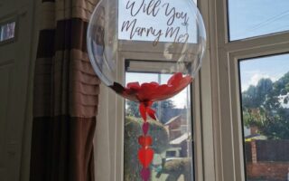 A clear balloon with "Will You Marry Me?" written on it floats indoors by a bay window. Red heart shapes are inside the balloon and strung in a line beneath it. Sunlight streams through the window, illuminating the scene.
