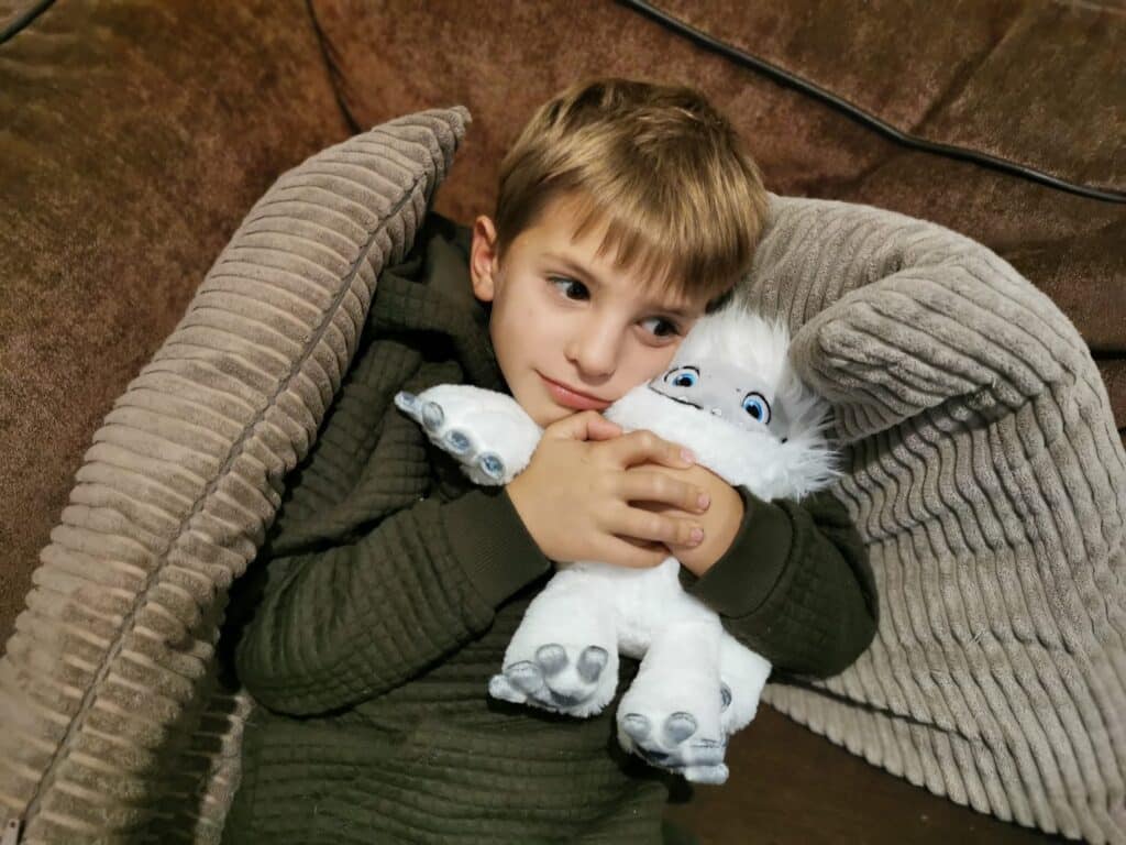 A young boy in a green jumper cuddles a fluffy white yeti soft toy while lying against soft, textured beige cushions on a brown sofa. He looks off to the side with a gentle, contented expression.