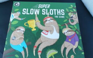A board game box titled "Super Slow Sloths: The Game" features four illustrated sloths in a race. One holds a "2021" trophy, another wears a medal, and two hold bananas. The background is green with leaves and a Venus flytrap. Slogan: "Get ready for sloths, puns and sabotage!".