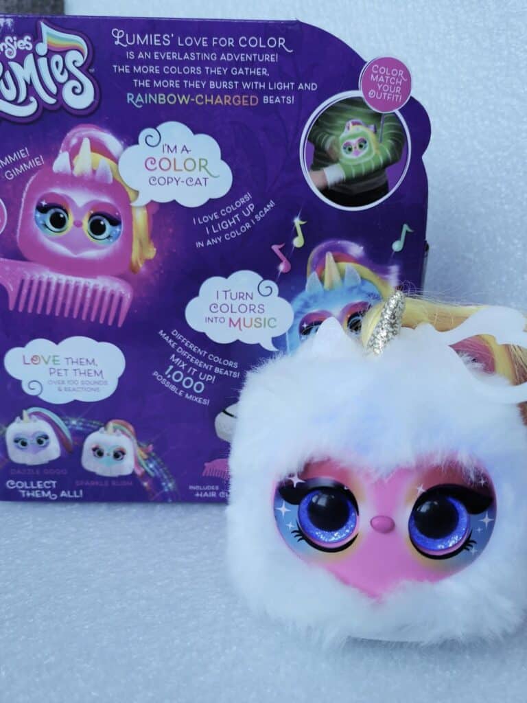 A soft toy with a white, fluffy body, pink face, large glittery eyes, and a gold unicorn horn is in front of its colourful packaging. The packaging shows another character and details features like light-up eyes and music colour change. A pink comb is included.