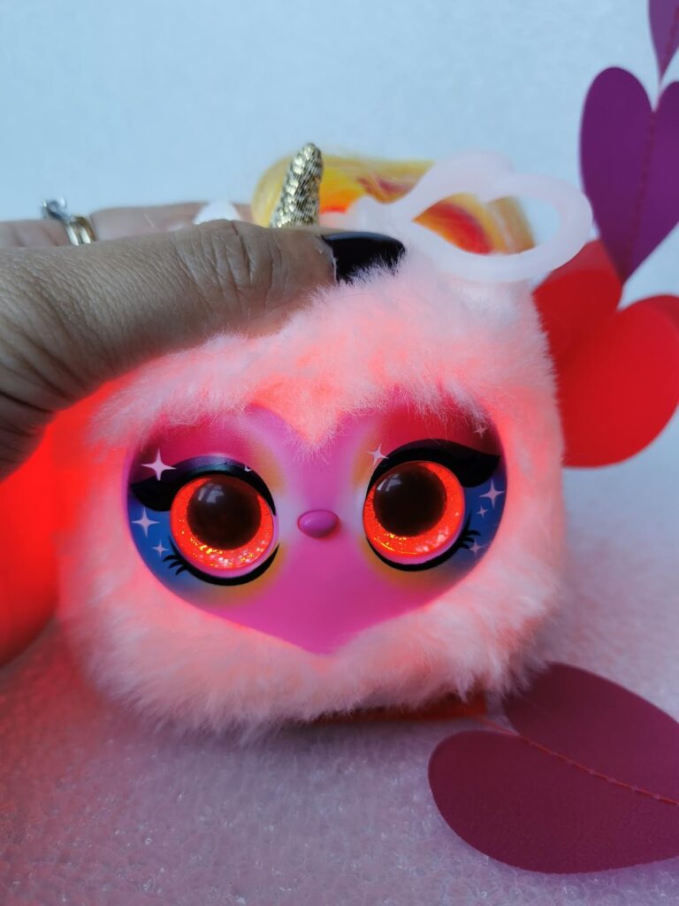 A hand holds a fluffy, round toy with glowing red eyes, a pink heart-shaped face, blue starry eyelids, and a small gold unicorn horn. Red and purple paper heart shapes are nearby on a white surface.