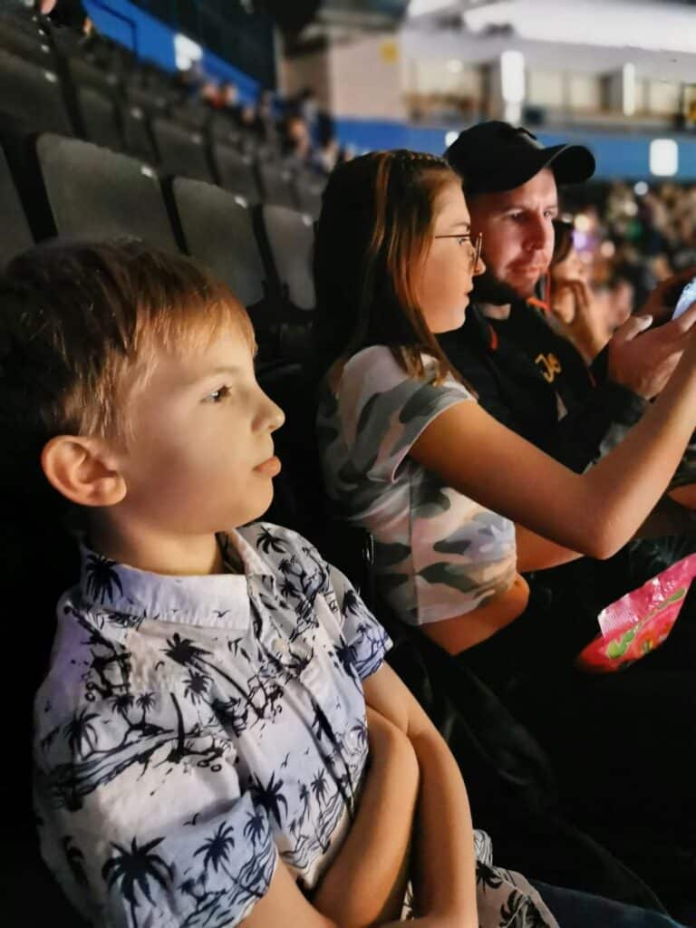 A young boy in a palm tree shirt sits with arms folded in stadium seats next to a woman in glasses and a man in a cap using a mobile. The background is filled with rows of empty black seats and a few blurred spectators.