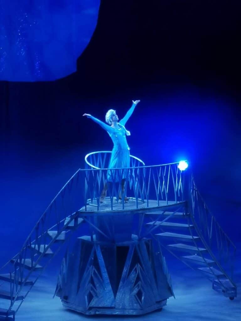 A performer dressed as Elsa from Frozen, in a sparkly blue gown and wig, stands on a raised staircase set with arms outstretched. Blue stage lighting and fog create a magical, icy atmosphere reminiscent of the “Let It Go” scene.