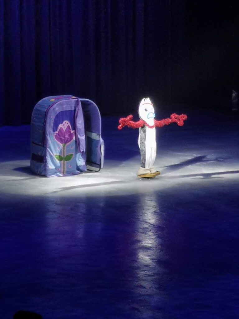 A person in a Forky costume with red pipe-cleaner arms stands on an ice rink stage next to a small, purple tent with a large flower design. The scene is lit by a spotlight, with dark blue curtains in the background.