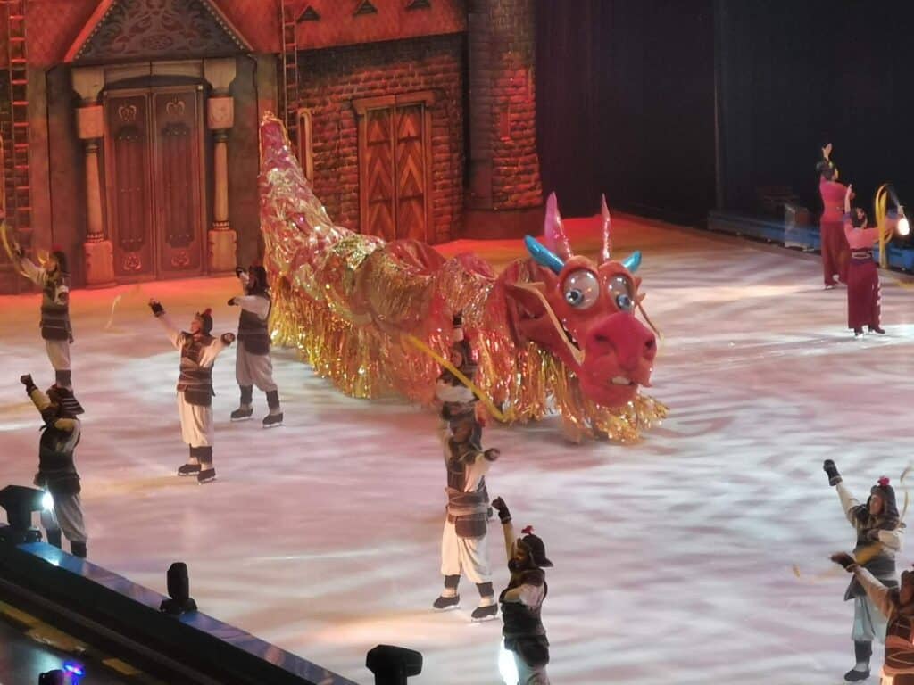Performers dressed in warrior costumes skate around a large, colourful dragon puppet with sparkly gold fringe on an ice rink. The set resembles an ornate, medieval-style building, and the stage is brightly lit, creating a festive atmosphere.