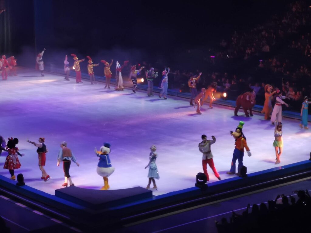 A group of Disney characters, including Mickey, Donald, and Goofy, perform on an ice rink in colourful costumes. They’re waving to an audience seated in a dark arena, illuminated by bright spotlights and purple stage lighting.