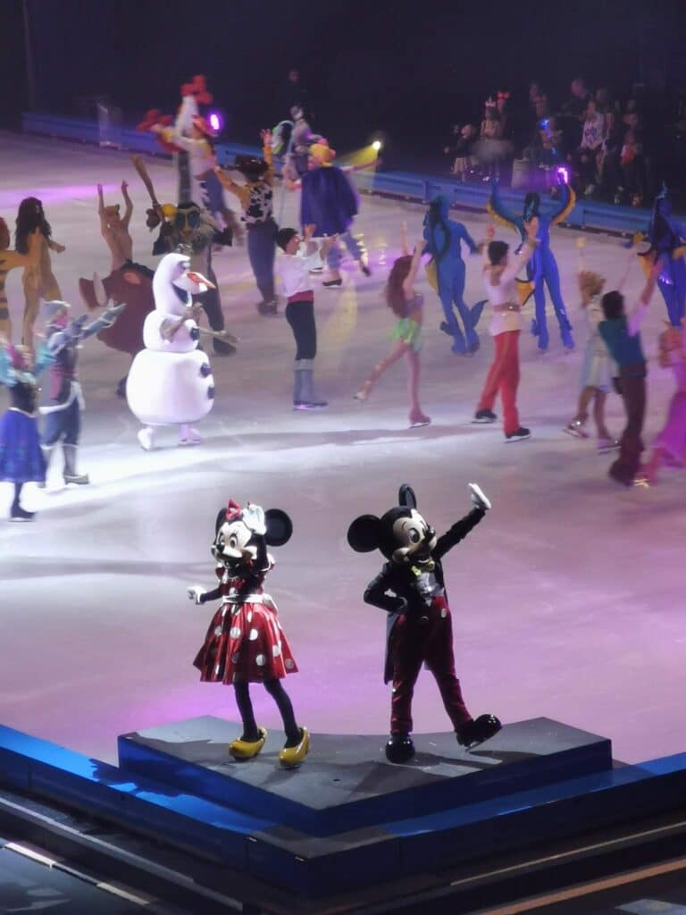 Mickey and Minnie Mouse stand on a platform, waving to the audience, as various Disney characters, including Olaf from Frozen, perform on an ice rink in the background under colourful stage lights.