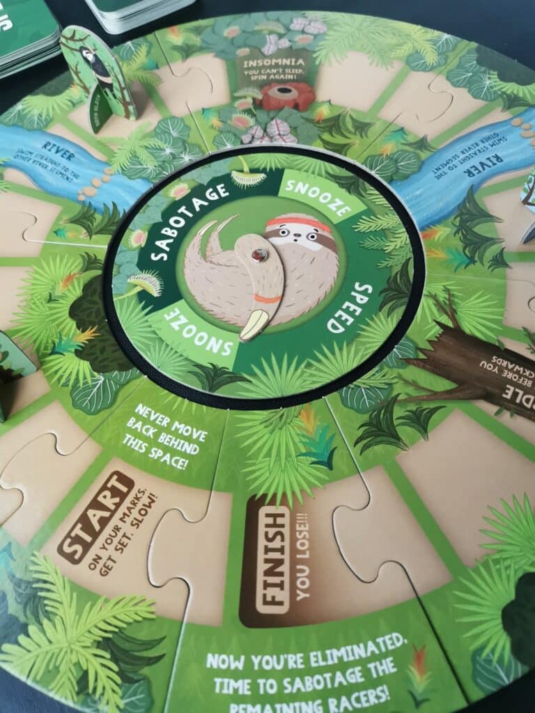 A circular sloth-themed board game features green jungle graphics and labelled sections like "Sabotage," "Snooze," "Speed," "Finish," and "Start." A sloth spinner is in the centre, with standing sloth character pieces and playful instructions on the game spaces.