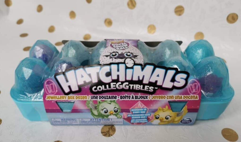 A light blue plastic egg box labelled “Hatchimals Colleggtibles” sits on a white surface with gold dots. The box has twelve sparkly blue eggs inside. Colourful cartoon creatures and branding are shown on the packaging’s front label.
