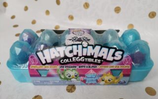 A light blue plastic egg box labelled “Hatchimals Colleggtibles” sits on a white surface with gold dots. The box has twelve sparkly blue eggs inside. Colourful cartoon creatures and branding are shown on the packaging’s front label.