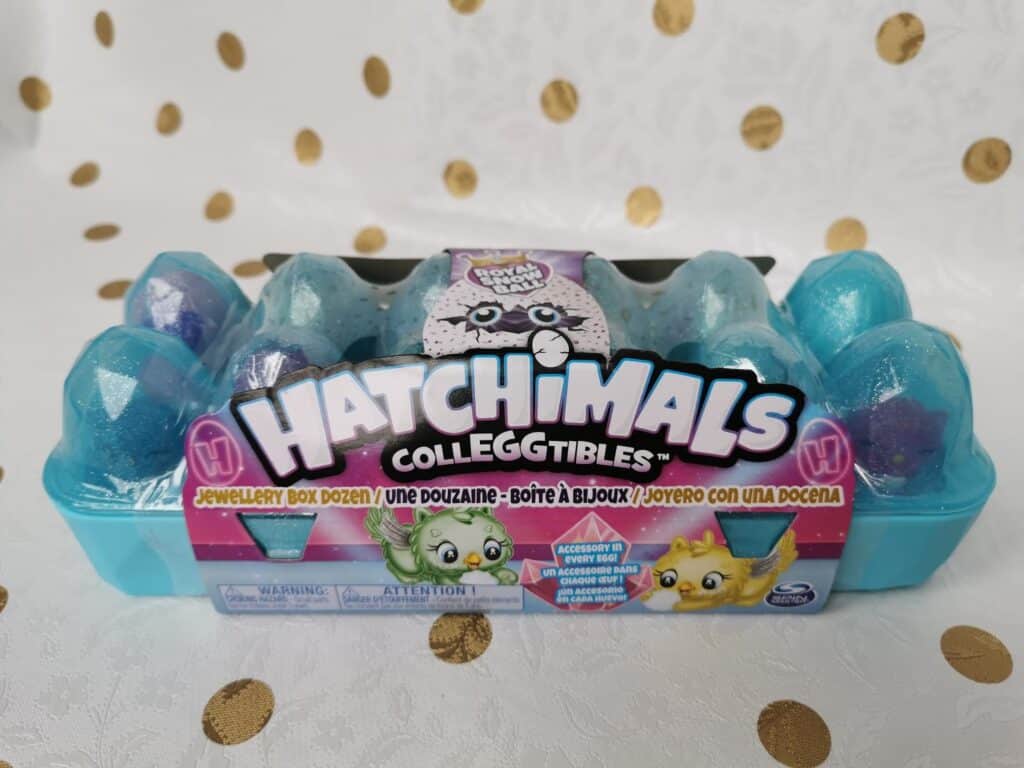 A turquoise plastic egg box labelled "Hatchimals CollEGGtibles" holds 12 speckled toy eggs. The packaging features cute illustrated creatures and colourful text. The background is a white cloth with gold polka dots.