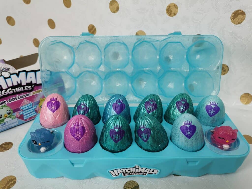 A blue plastic egg carton holds ten colourful toy eggs, mostly teal, some pink, each with a purple heart. Two small animal figurines, one blue and one pink, are placed in front. "Hatchimals Colleggtibles" logo is on the carton. Gold polka dot background.