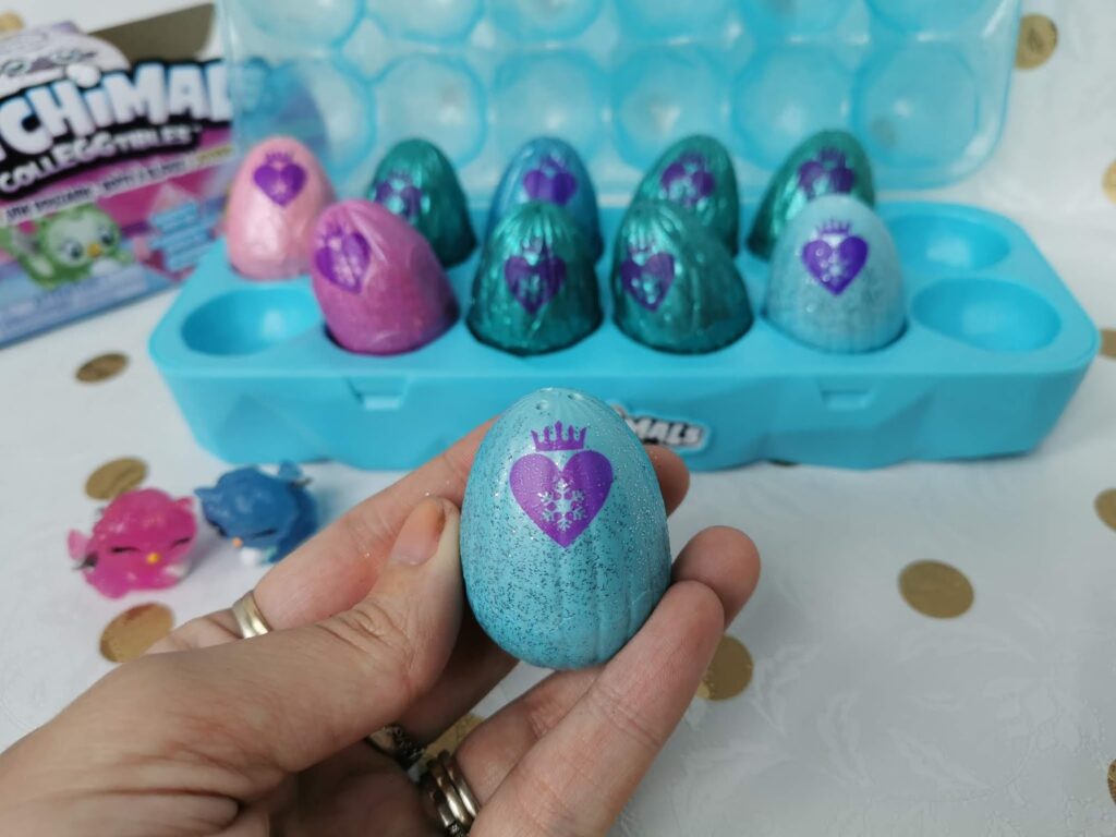 A hand holds a blue speckled egg with a purple heart and crown printed on it. In the background, more colourful eggs are arranged in a blue plastic egg tray, with toy figures and packaging partially visible.